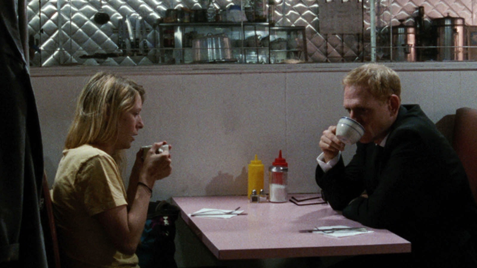 Film still from Slow Machine. A young woman sits nervously at a diner table with a man. Both of them are sipping coffee and tea.