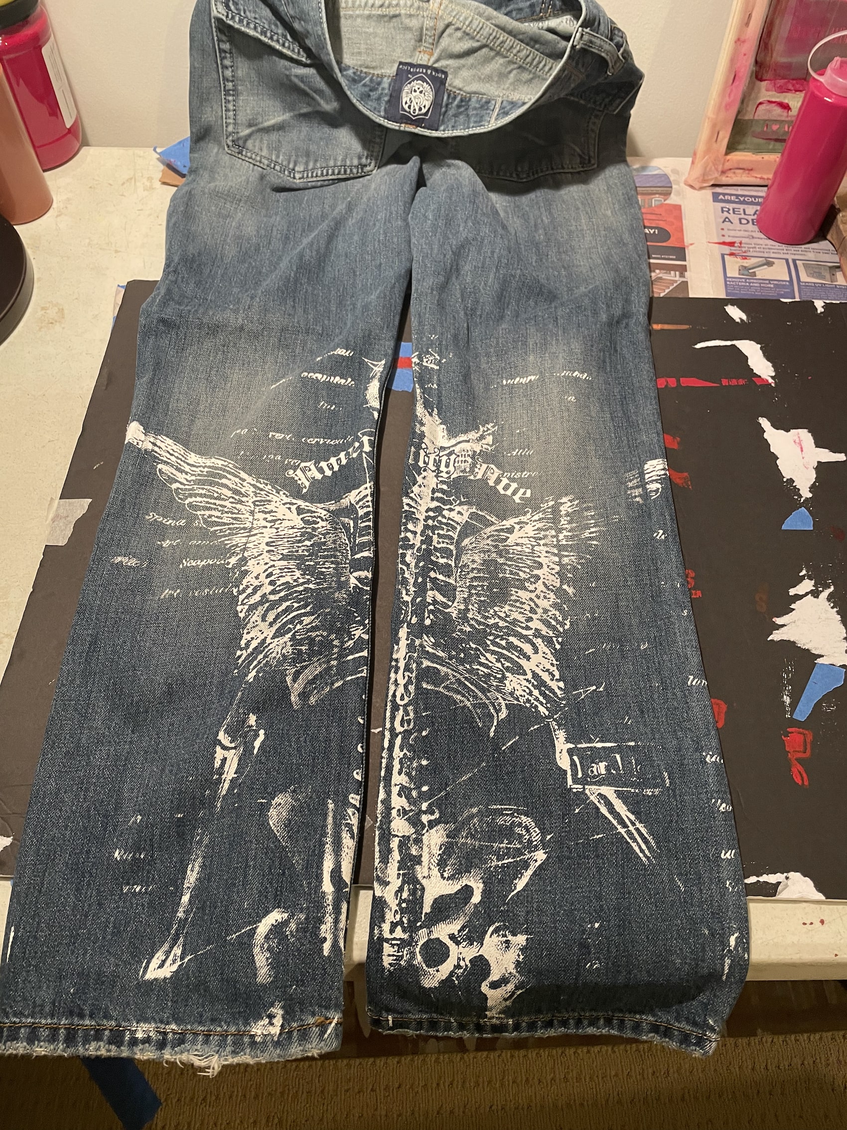 still image of a thrifted pair of jeans with fairy skeleton design screen printed in white ink laying face-down on a table