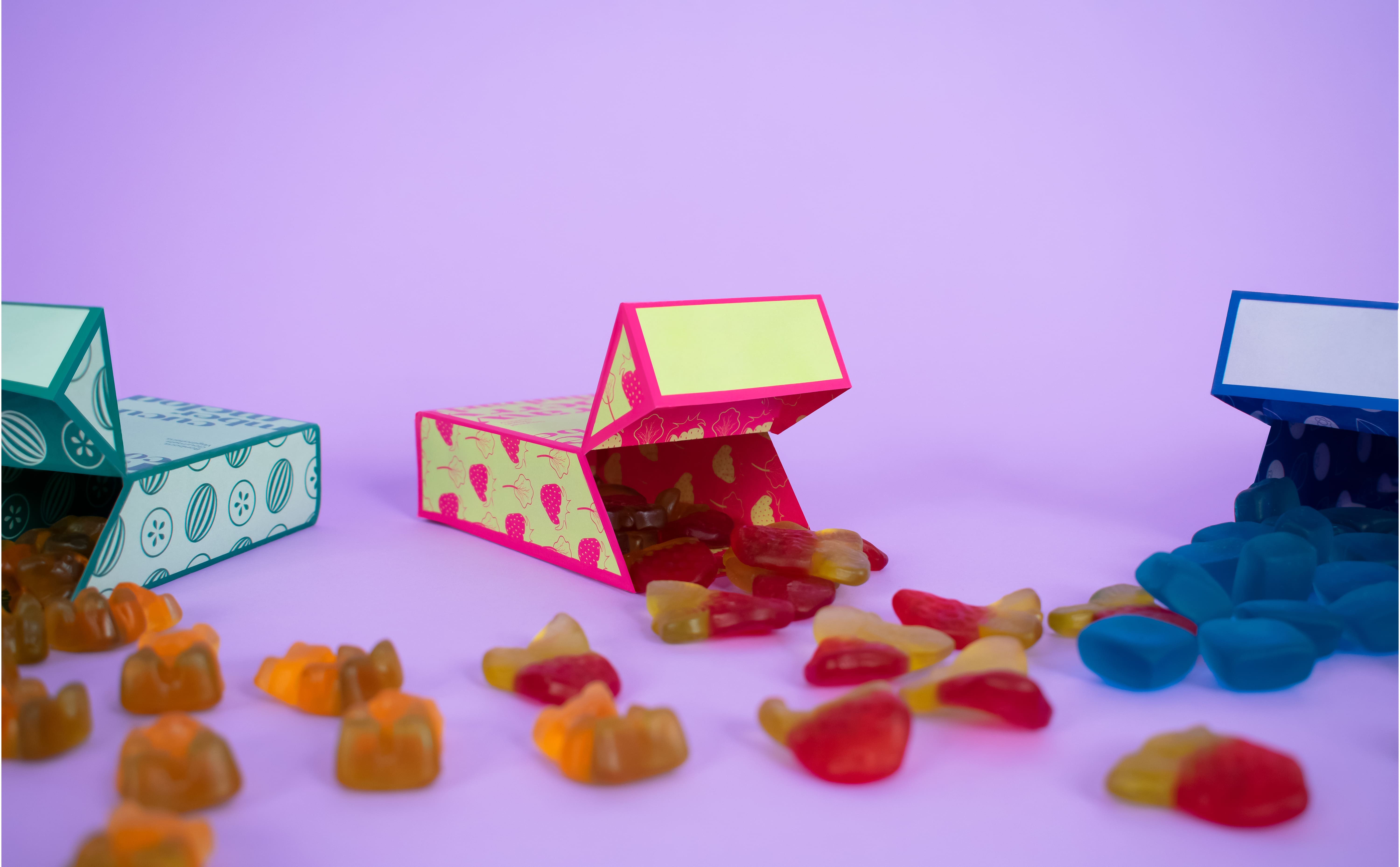 A trio of colorful packages with open lids. There is a mix of gummy candies everywhere on a purple background.