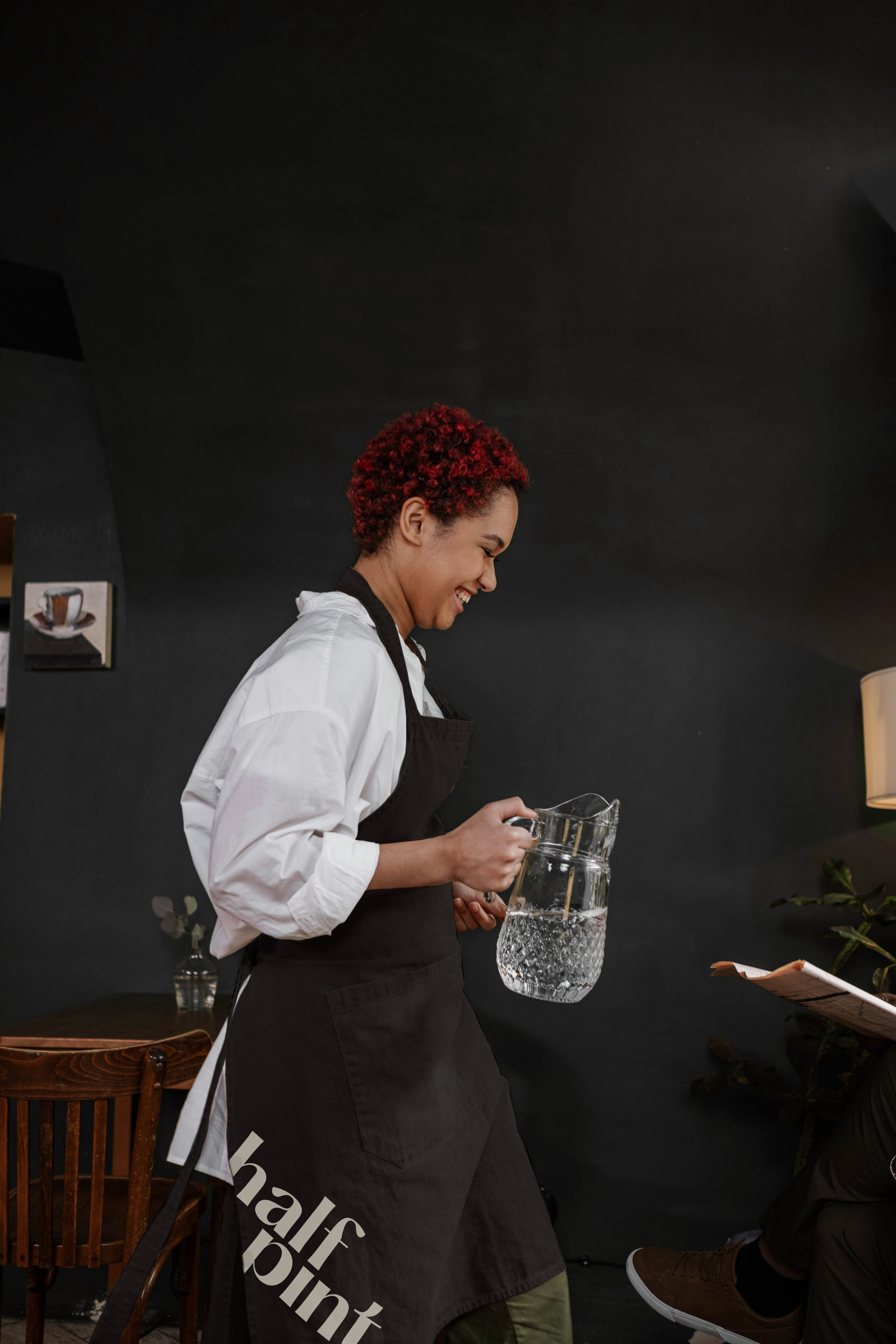 A waitress wearing a black apron with a logo in beige at the bottom of it. She is walking and carrying a jug of water.