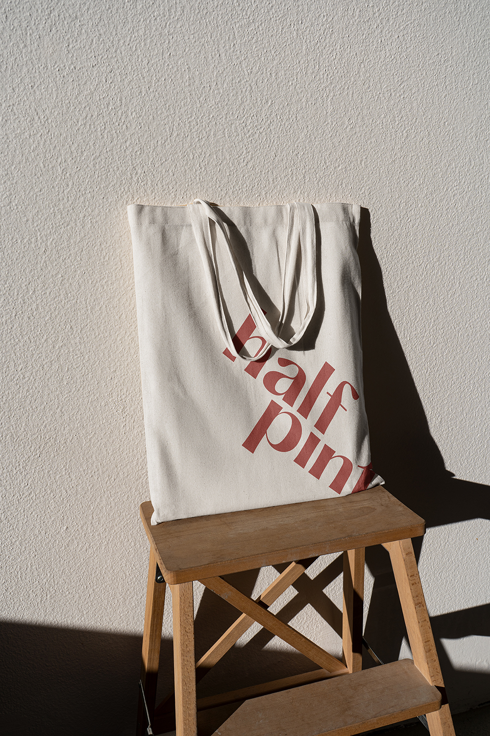 A beige canvas tote bag with red writing. The bag is on a wooden stool and leaning on a concrete wall.