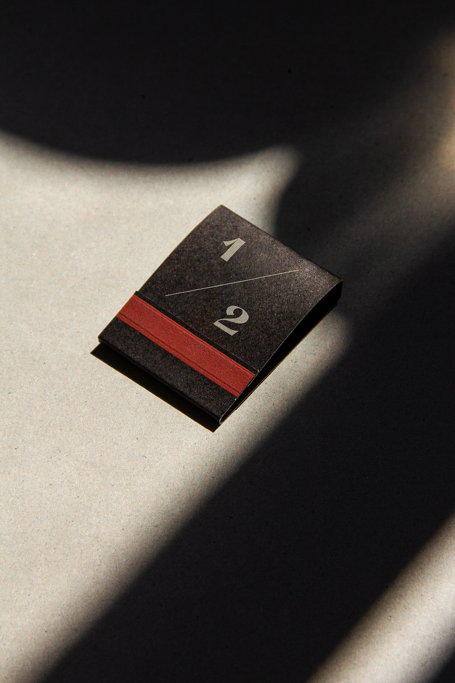 A black matte pack of matches with a red bar and a half fraction in beige. The matches are lying on a concrete surface.