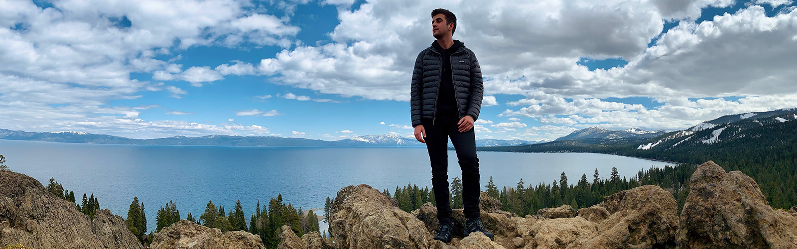 Charles Holeman standing on a rocky surface. The background is a beautiful lake contrasting with a snowy mountain range and sunny but slightly cloudy sky. 