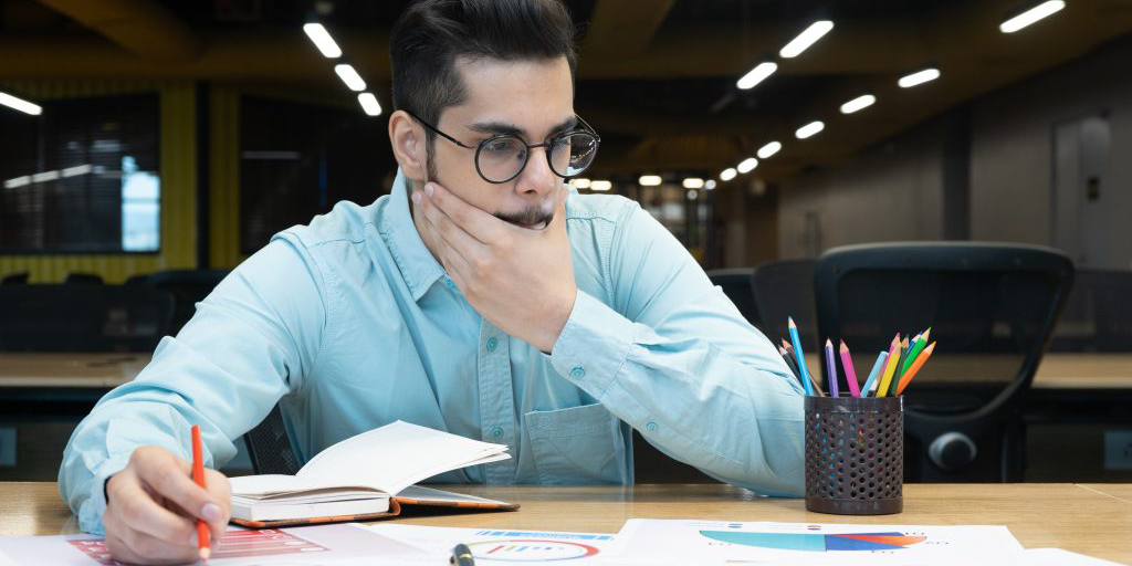 Male bearded office worker at a desk facing the camera, looking to the bottom right corner of the image with his hand over his mouth. Looks to be deep in thought. He is holding a pencil in one hand.