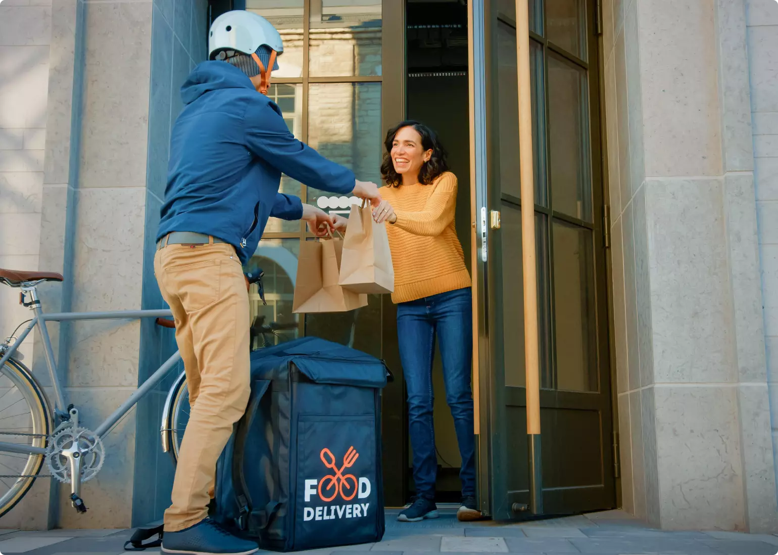 A smiling young woman with shoulder-length dark hair stands in the open doorway of an apartment building. A man in a cycling helmet, whose face we cannot see, is handing her two unmarked brown paper bags. An insulated backpack marked "Food delivery" is on the ground at his feet.