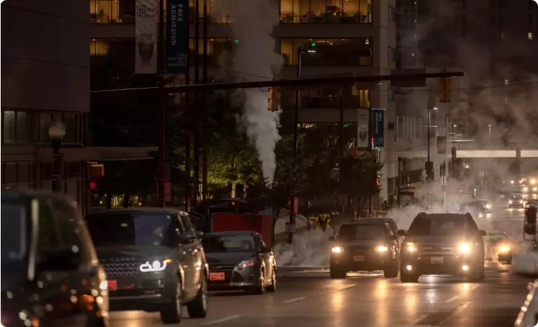 Nighttime view of a busy road in Baltimore, Maryland. Lights are visible in tall buildings on either side of the road, and there is a cloud of mist or steam over the road.