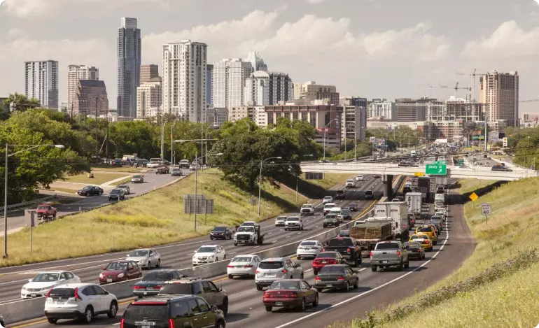 A view of a packed highway leading into downtown Austin, Texas. There are tall buildings in the background and the highway is flanked by grassy banks.