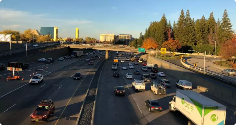 A busy freeway in Sacramento, California. There is an overpass in the background, with some tall fir trees behind it.