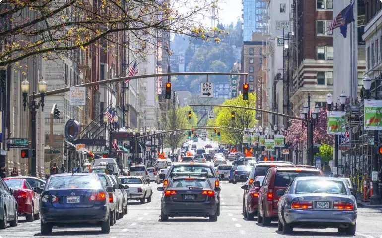 Busy urban street in Portland, Oregon. The street is lined with tall buildings and trees showing blossoms or fresh spring leaves. The road is dominated by car traffic.