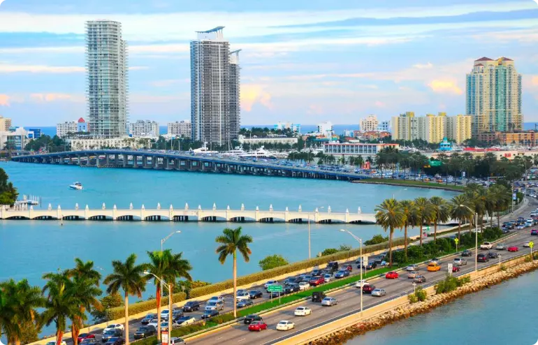Aerial view of Miami, Florida. A palm-lined road in the foreground curves around a blue-water bay towards the city with tall towers in the background.