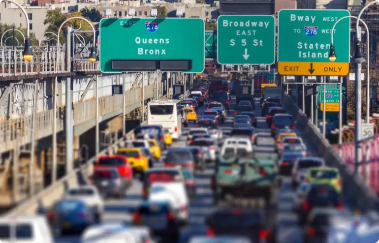 A clogged highway in New York. Signs across the road read, from left to right: "Queens, Bronx"; "Broadway East"; and "Bway West to 278 West, Staten Island, Exit Only".