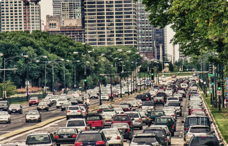 An urban road in Chicago is choked with traffic across four lanes in both directions. The road is lined with grass and tall trees, and tall buildings are visible in the background.