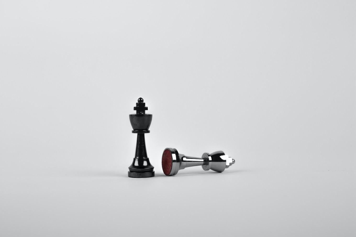 A black chess king standing next to a fallen silver chess king on plain white background