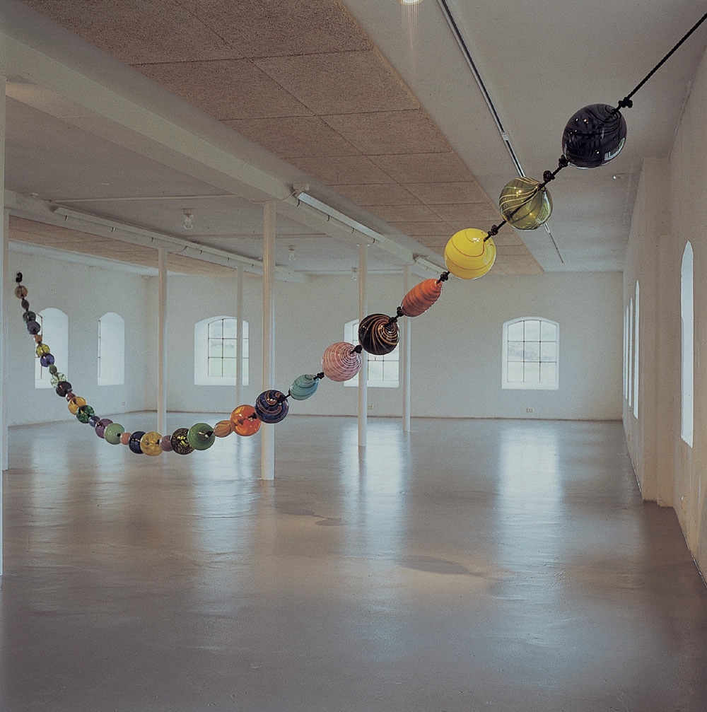 A colossal blown glass bead necklace by Michael Petry hung in an art museum  