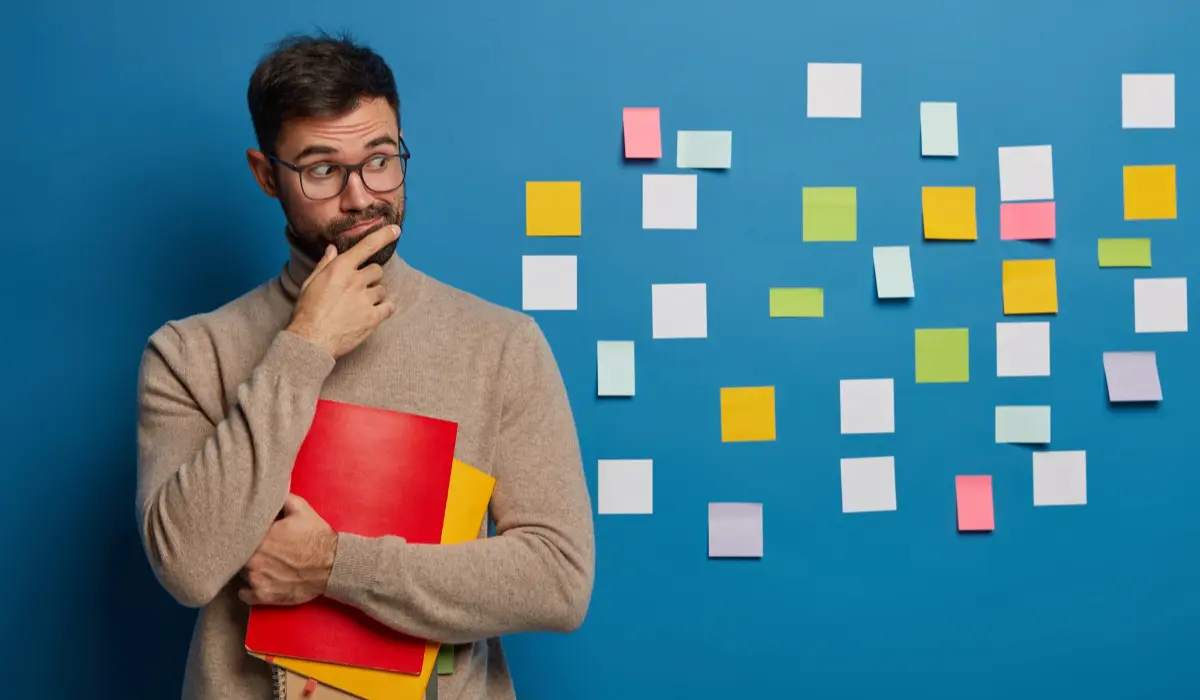 a man holding files in hand and thinking in front of a blue wall with sticky notes on it