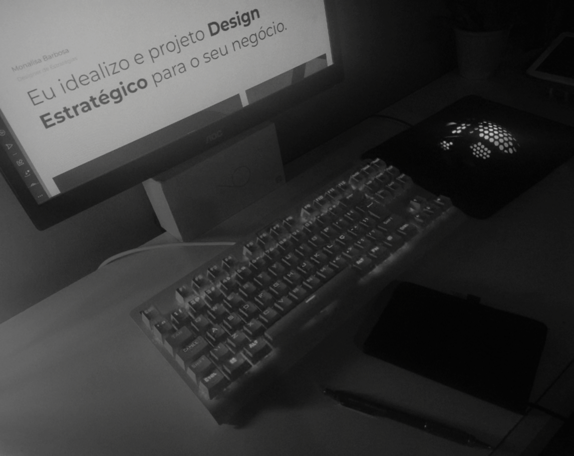 Foto em Preto e Branco de uma mesa de trabalho, composta pela tela, mouse, teclado, caderno de anotações fechado e uma lapiseira