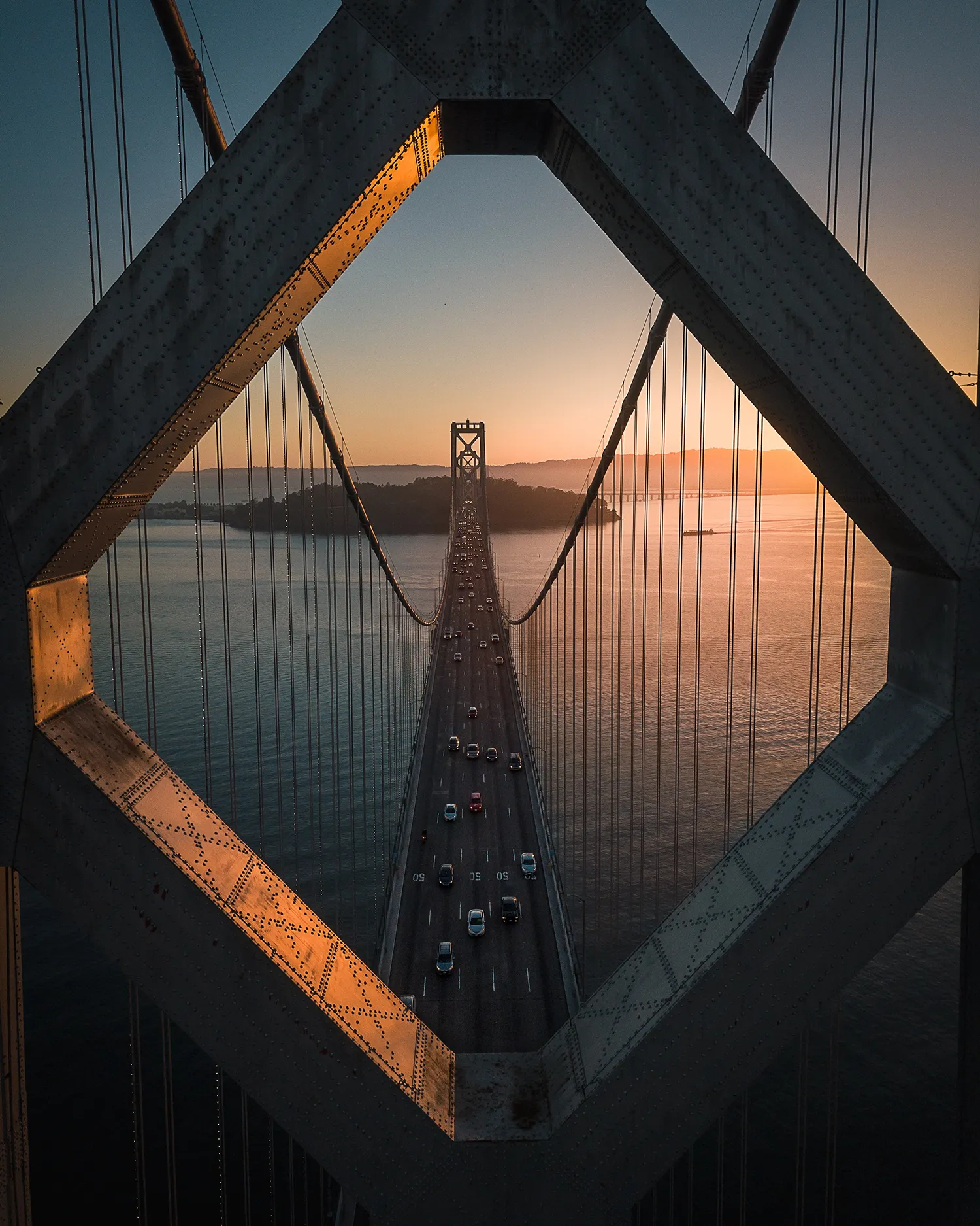 A view of the sunset can be seen through the Bay Bridge in California, with light shimmering across the water below.