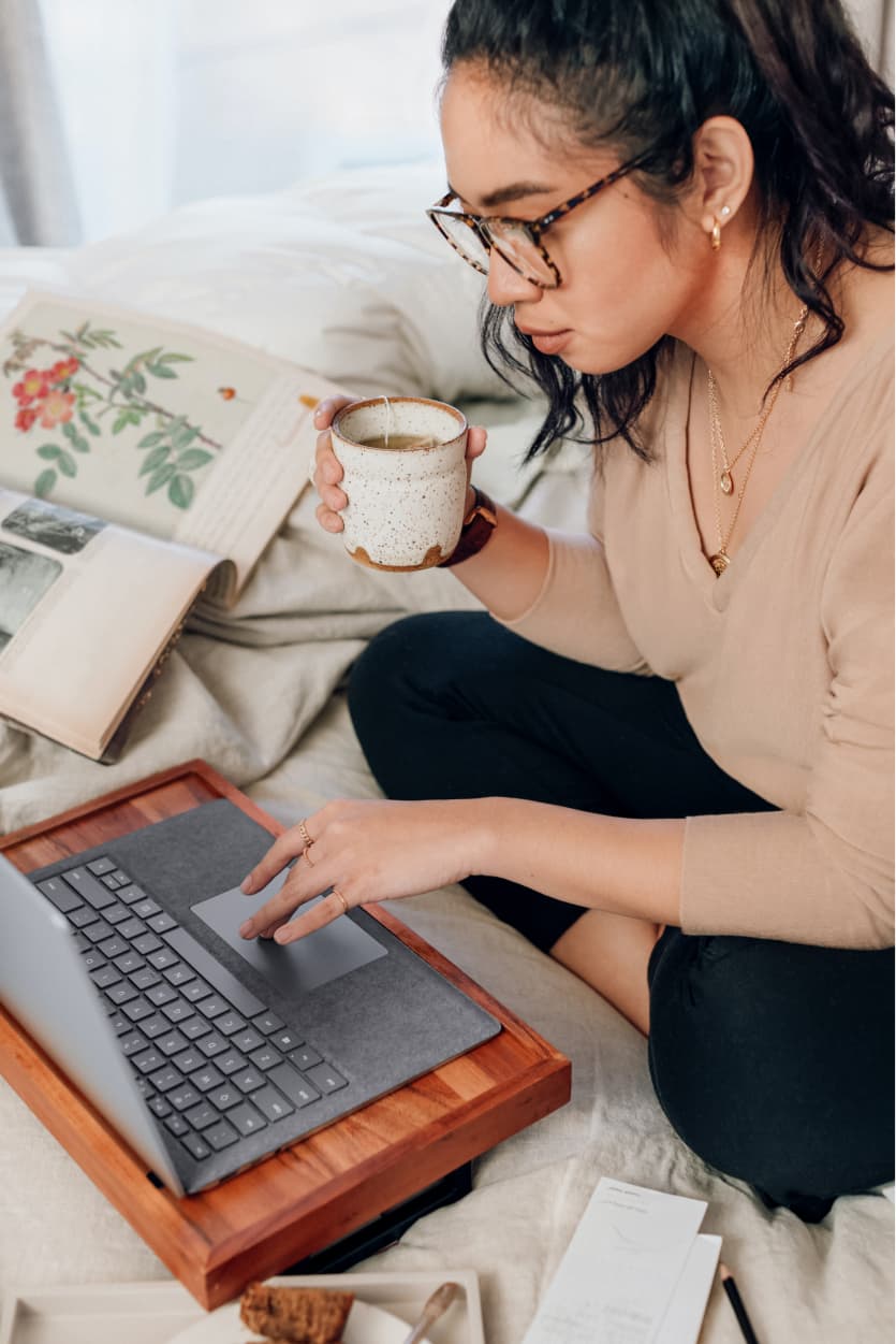 A 30-something asian woman holding a cup of tea, typing on a laptop sitting on a cozy bed with an open book next to her.