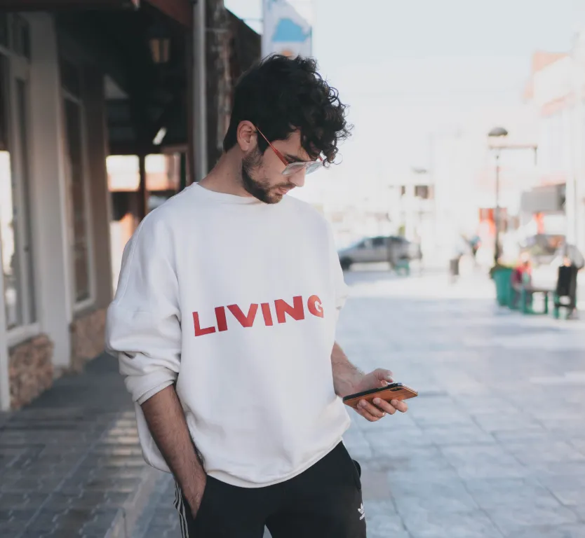 a 20-something year old man standing in the street looking down at his mobile phone. he is were a white sweatshirt with the word "living" printed in red on it.