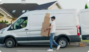 An unmarked white delivery van is parked on a suburban street with its rear doors open. A young man in jeans, white shirt and a black baseball cap is carrying a stack of four plain brown cardboard boxes.