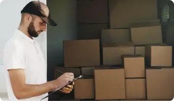 A man in a white shirt and brown baseball cap makes a note on a clipboard. He is standing at the open door of a delivery van full of plain, unbranded cardboard boxes.