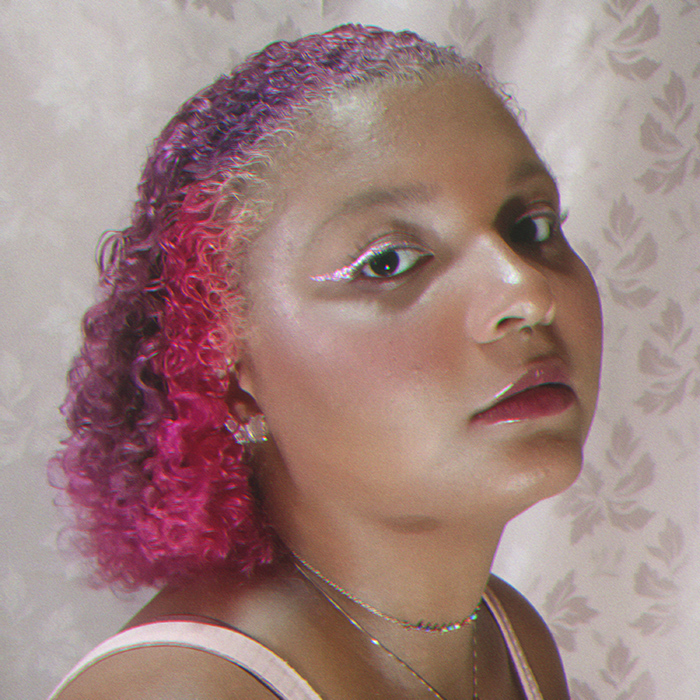 sarah borrete de lado, olhando para a câmera, num fundo rosa