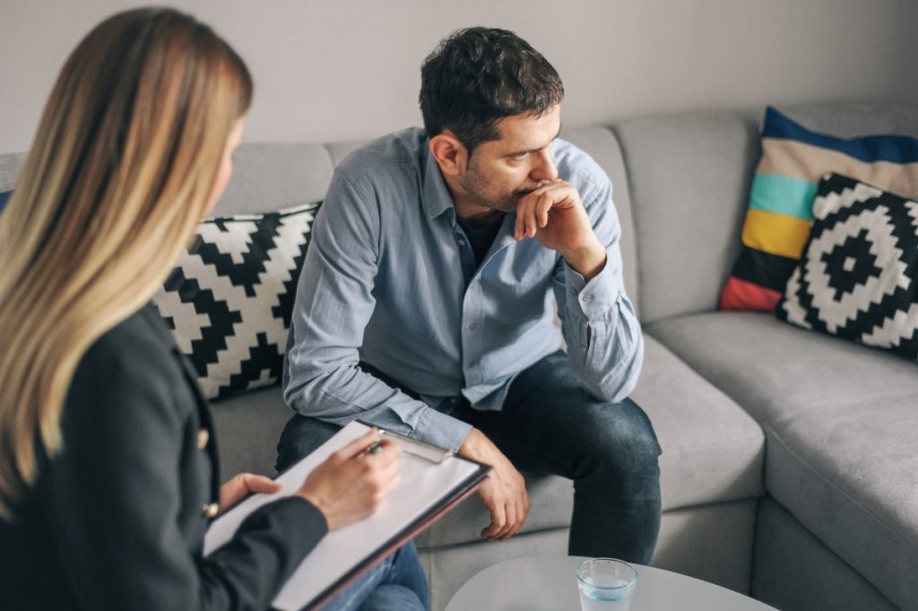 A young bearded man in a blue button-down shirt sits on a couch and reports an instance of sexual harassment. He is looking away from the woman who is taking notes of his complaint.