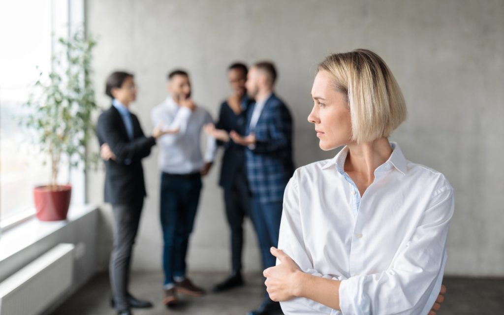 A woman in white blouse stands uncomfortably in the foreground as a group of men in the background make comments about her.