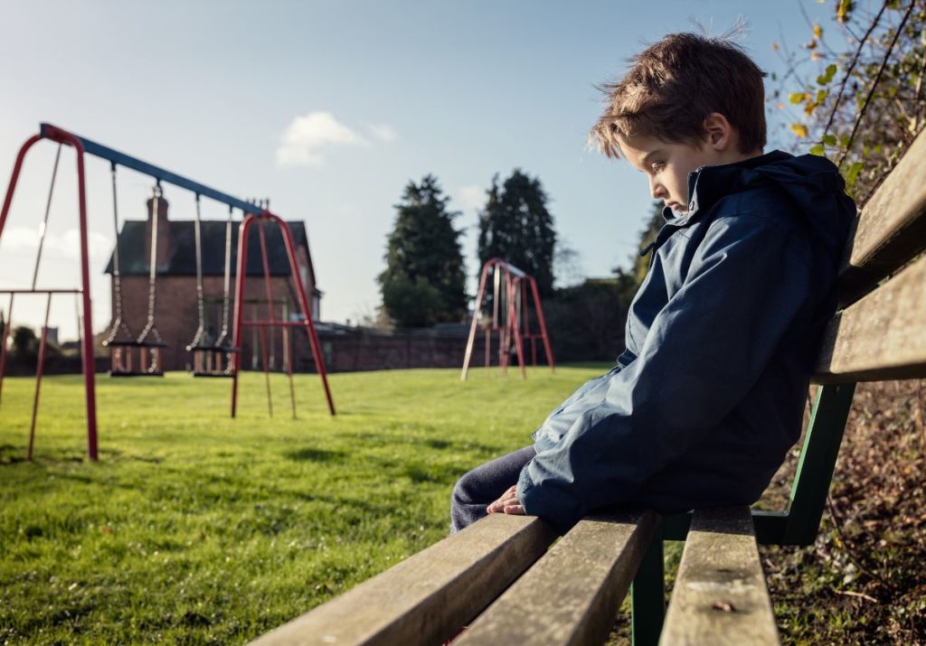 A boy in a blue jacket sits alone beside a swing set looking sadly at the ground.