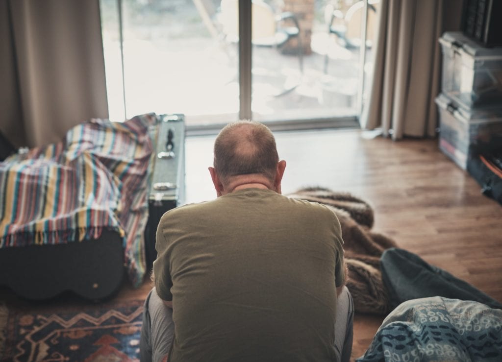 Middle-age man sitting with back to the camera in bedroom with glass sliding door