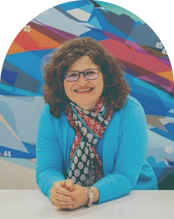 A photo of Wendy Thomas. Wendy is a white woman with shoulder-length, brown, curly hair and rectangular glasses frames. She is leaning forward with her elbows resting on a counter. She is wearing a bright blue sweater and polka dot scarf, against a blue and orange colored backdrop.