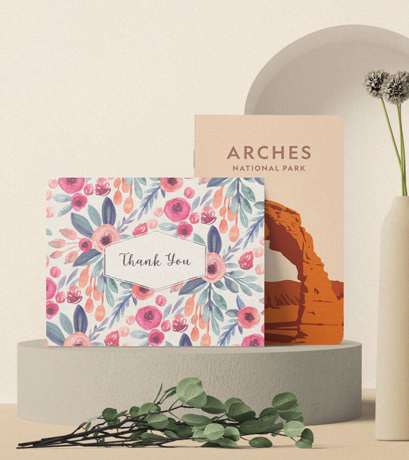 A thank you greeting card and arches national park notebook placed on a round cement with a branch with green leaves in front of it.