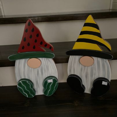 Two garden gnomes with long bears. One is wearing a hat that looks like a water melon slice and shoes that look like watermelon rinds. The other is wearing a hat decorated like a bumble bee and black shoes. 
