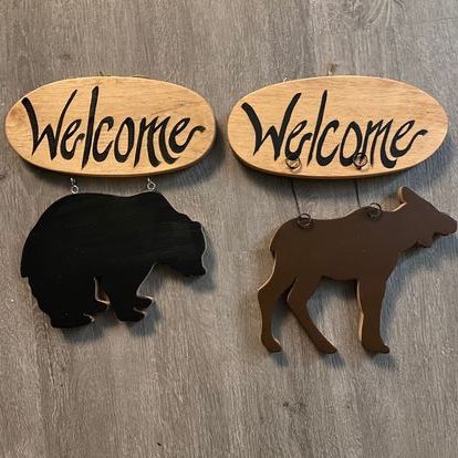 Two Signs that say "welcome". One decorated with a black bear, the other with a moose.