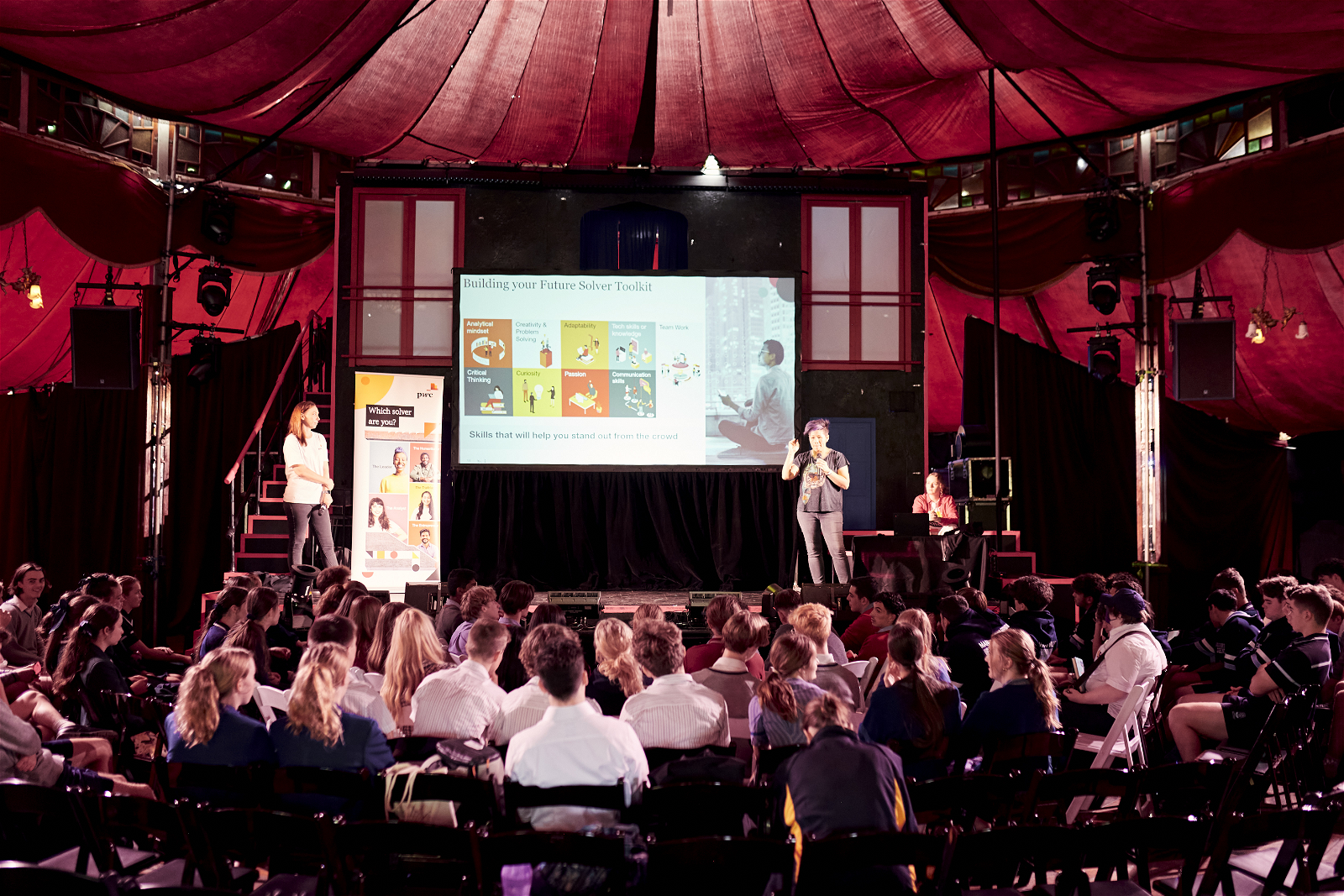 Two female speakers on stage in front of a large presentation screen, presenting to a large audience of students.