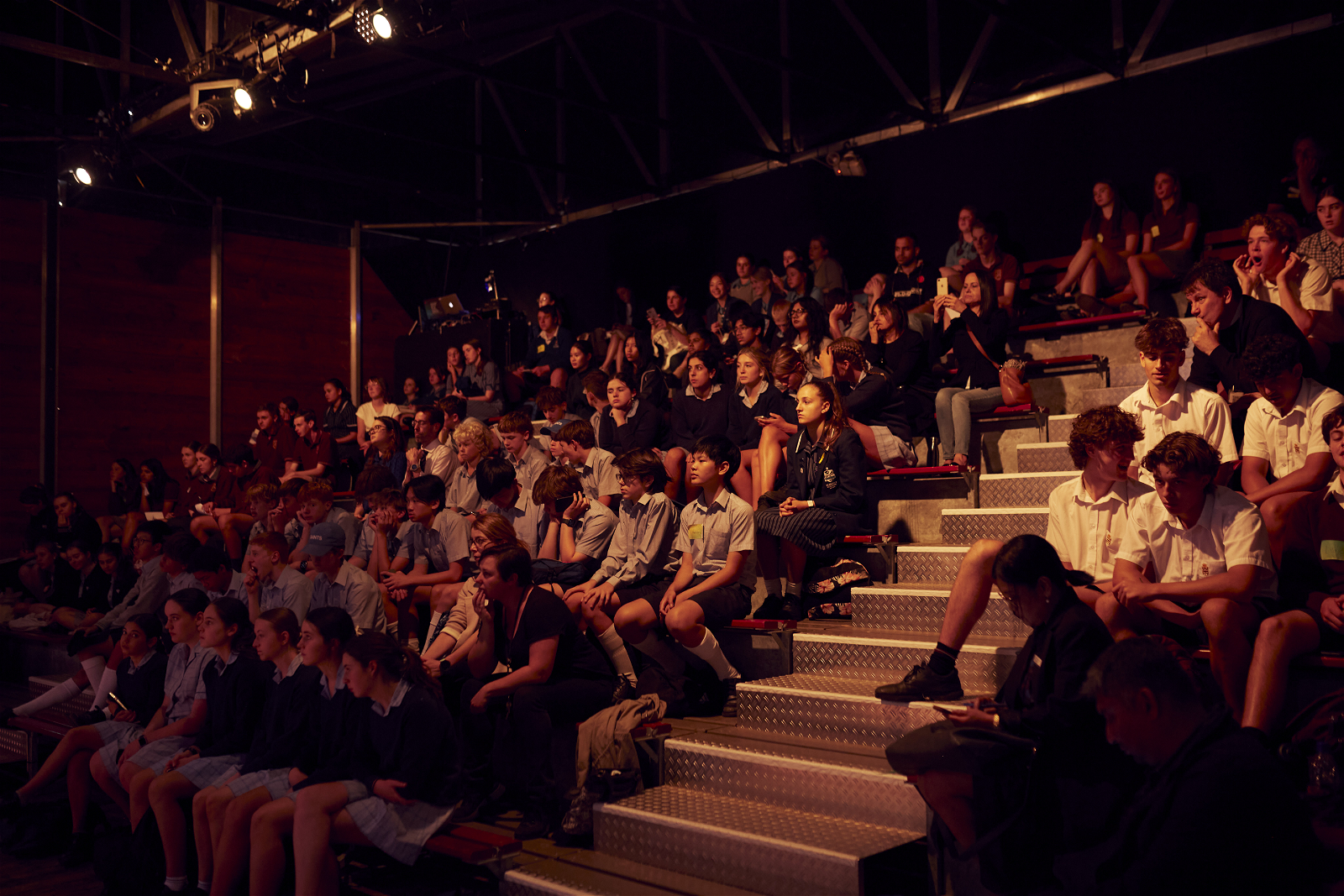 A large group of students sitting on bleachers inside a Fringe venue in low lighting, watching a speaker from the Startup Academy line-up.