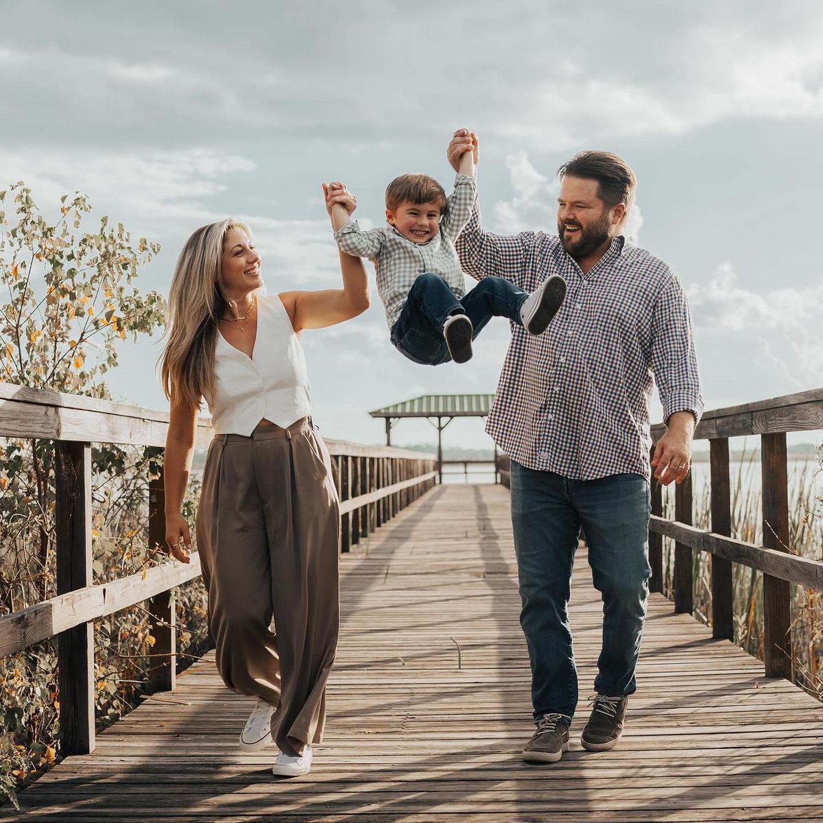 Unique Wedding Photography Ideas - Joyful family moment with parents swinging their child between them on a sunny wooden boardwalk, surrounded by nature.