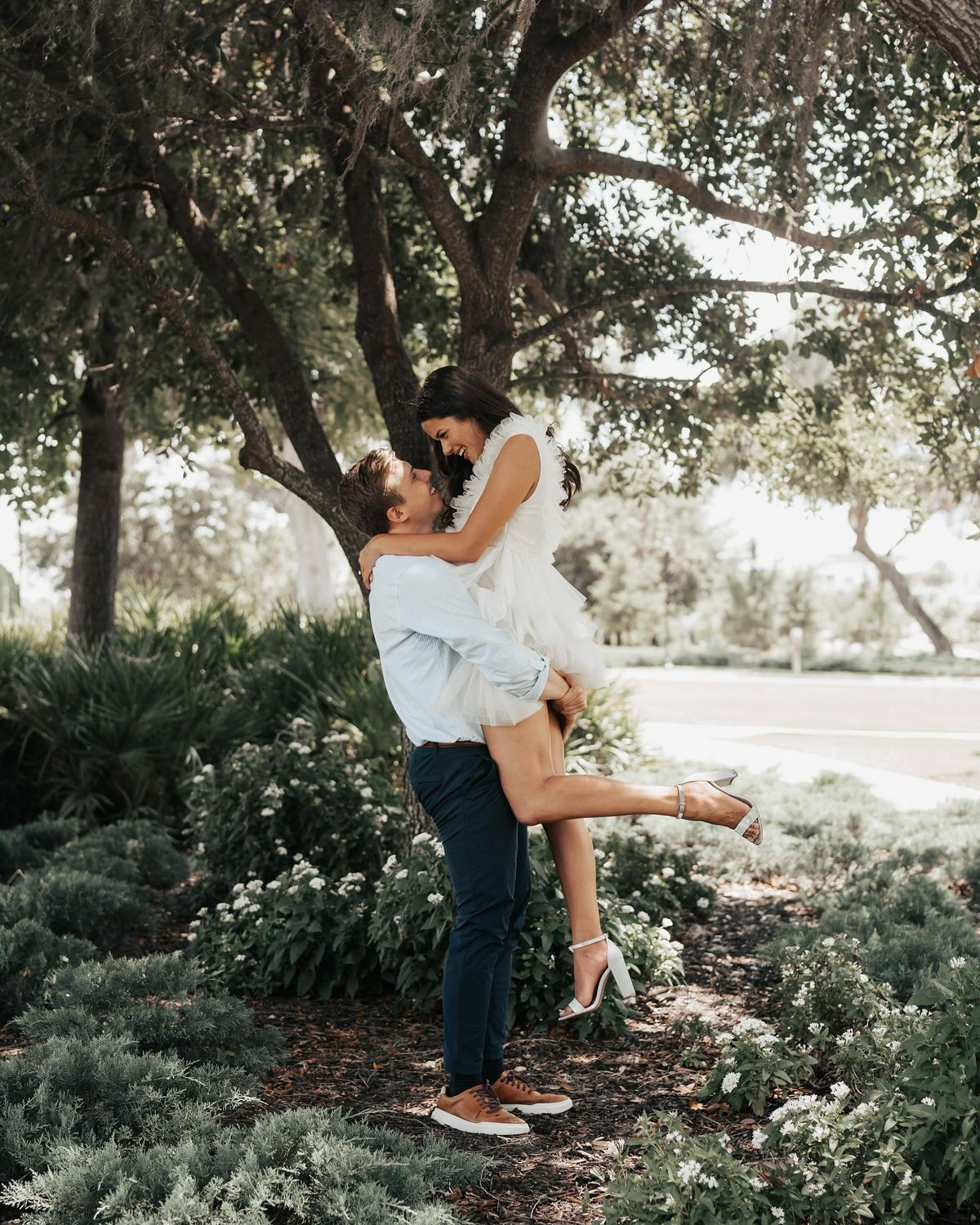 Cinematic Wedding Video - A man lifting a woman in a joyous embrace under a shaded tree, surrounded by lush greenery, capturing a candid moment of love and happiness.