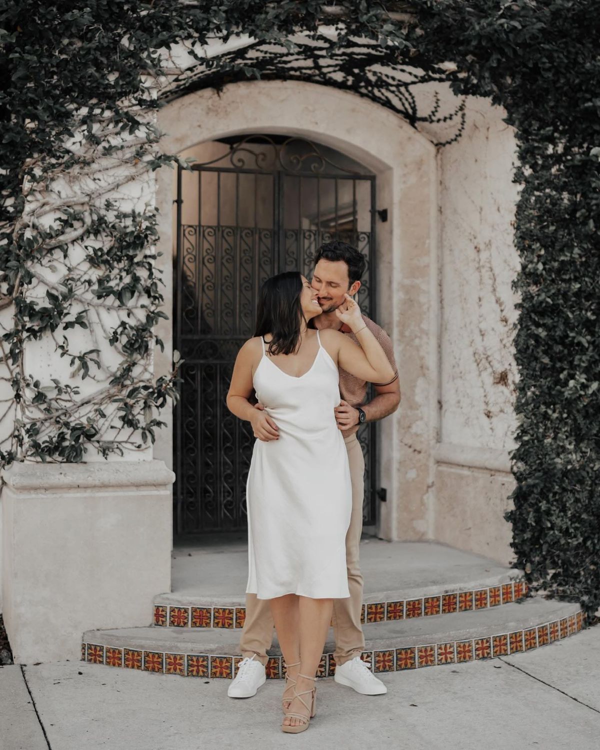 How to Create a Wedding Timeline - A charming couple sharing an intimate moment, the man whispers into the woman's ear, making her smile, framed by lush greenery and a vintage doorway.