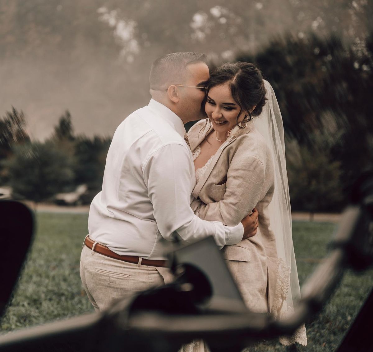 Hacienda Siesta Alegre in Puerto Rico - A bride and groom sharing a tender moment, with the bride smiling joyfully as the groom whispers in her ear, amidst a serene setting with a vague camera foreground hinting at a captured moment.
