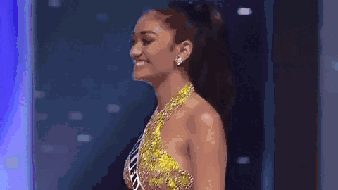 A woman in a sparkling yellow gown confidently walks on stage, smiling and waving to the audience. The background has blue and purple lights, adding to the glamorous atmosphere. She exudes confidence and grace as she makes her entrance.