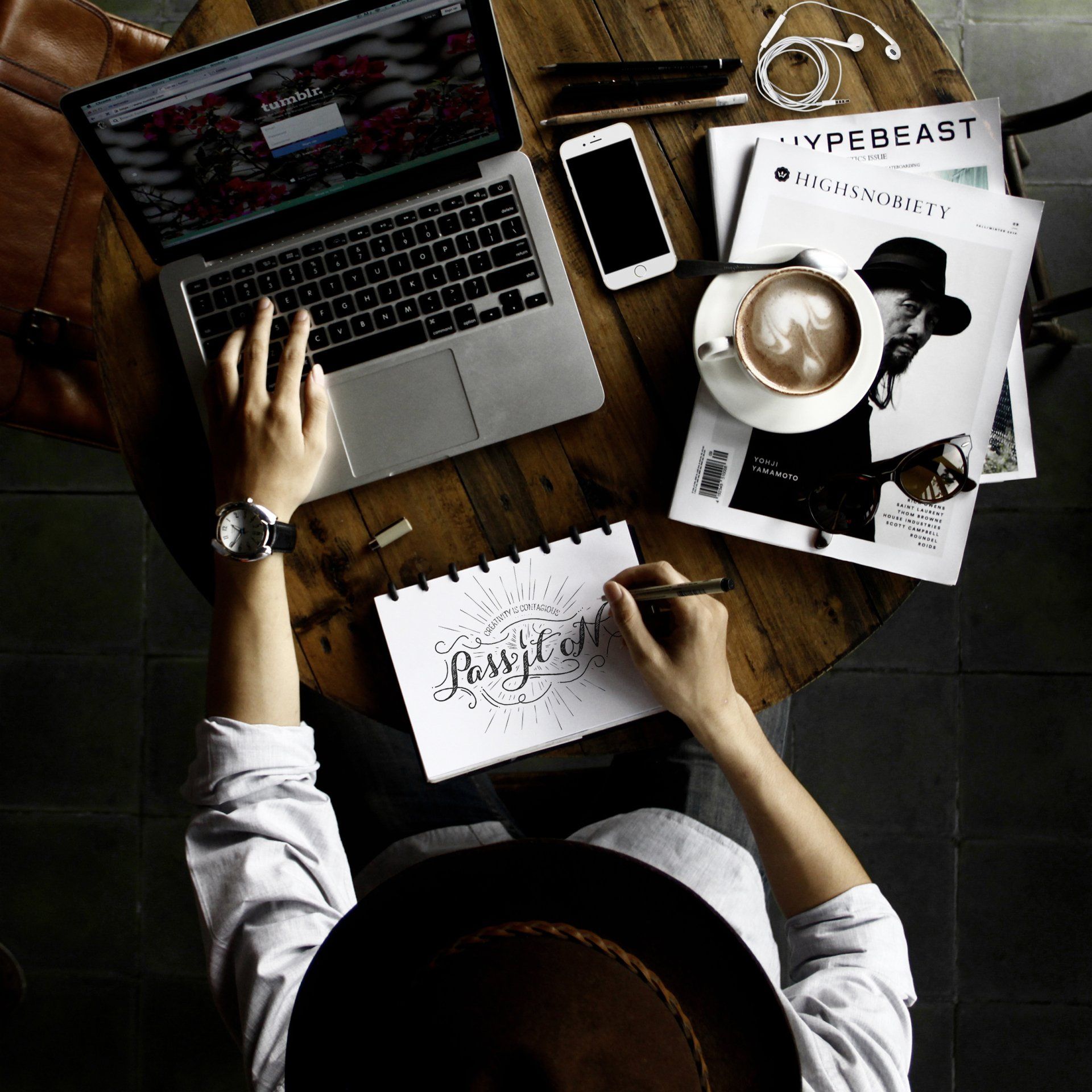 Creative leader working on laptop while sketching typography design, with coffee, smartphone, magazines, and workspace tools on rustic wooden desk.