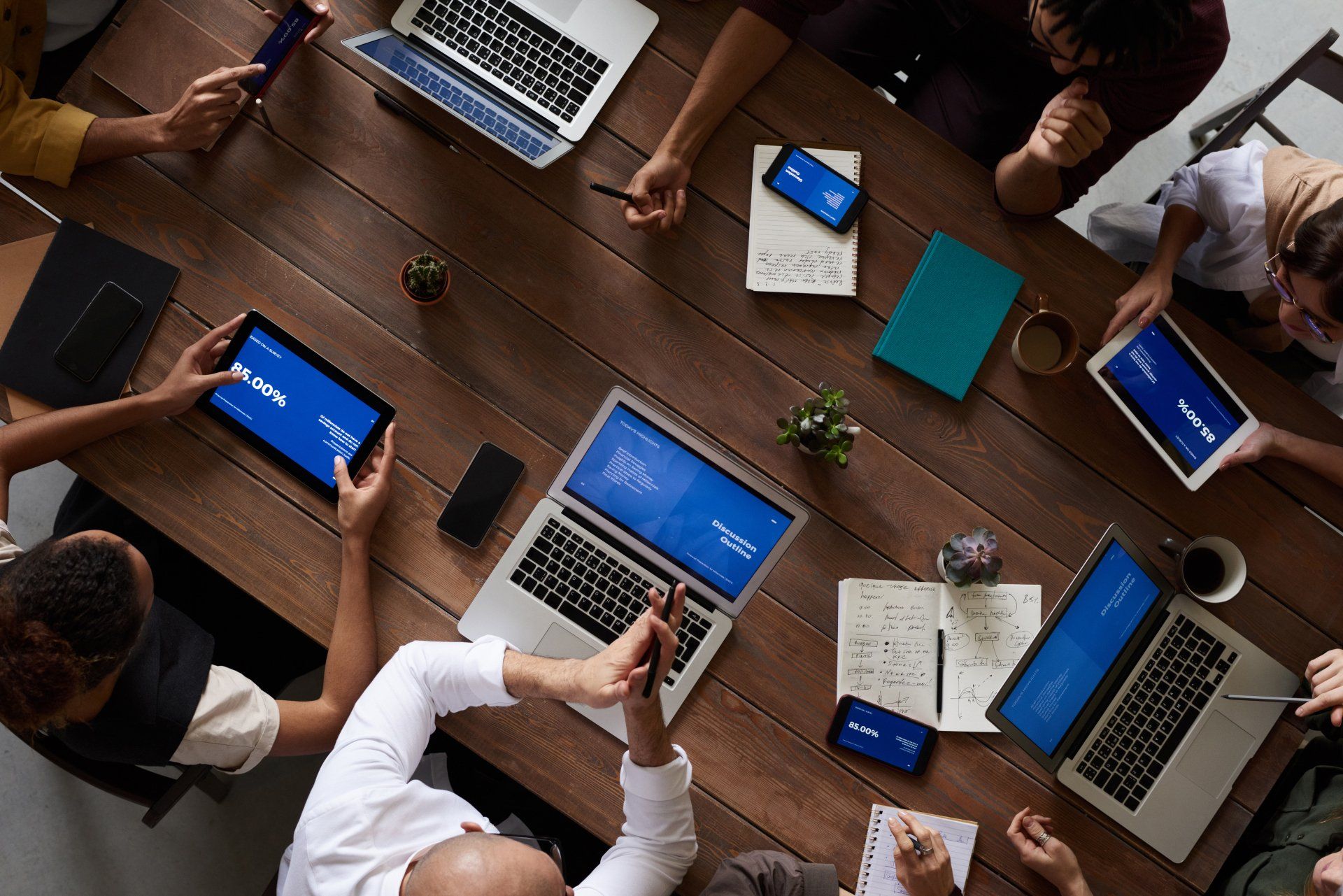 Sales leaders and team collaborating around a wooden table with laptops, tablets, and smartphones displaying sales performance data and metrics, discussing sales growth strategies.