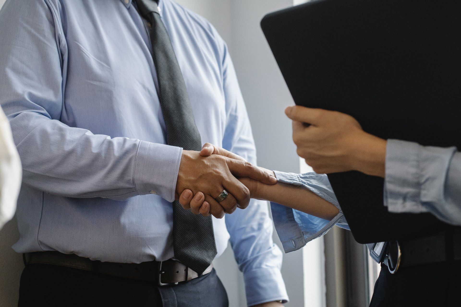 Two business professionals shaking hands during a successful sales negotiation, with a third person holding a clipboard in a corporate office setting.